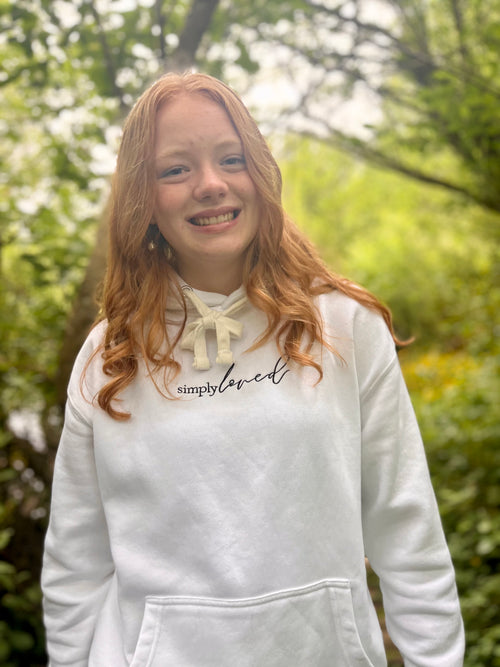 Girl smiling in a white “simply loved” hoodie, standing in a soft, green forest setting. Minimalist faith-based apparel by Simply Sofia Joy.