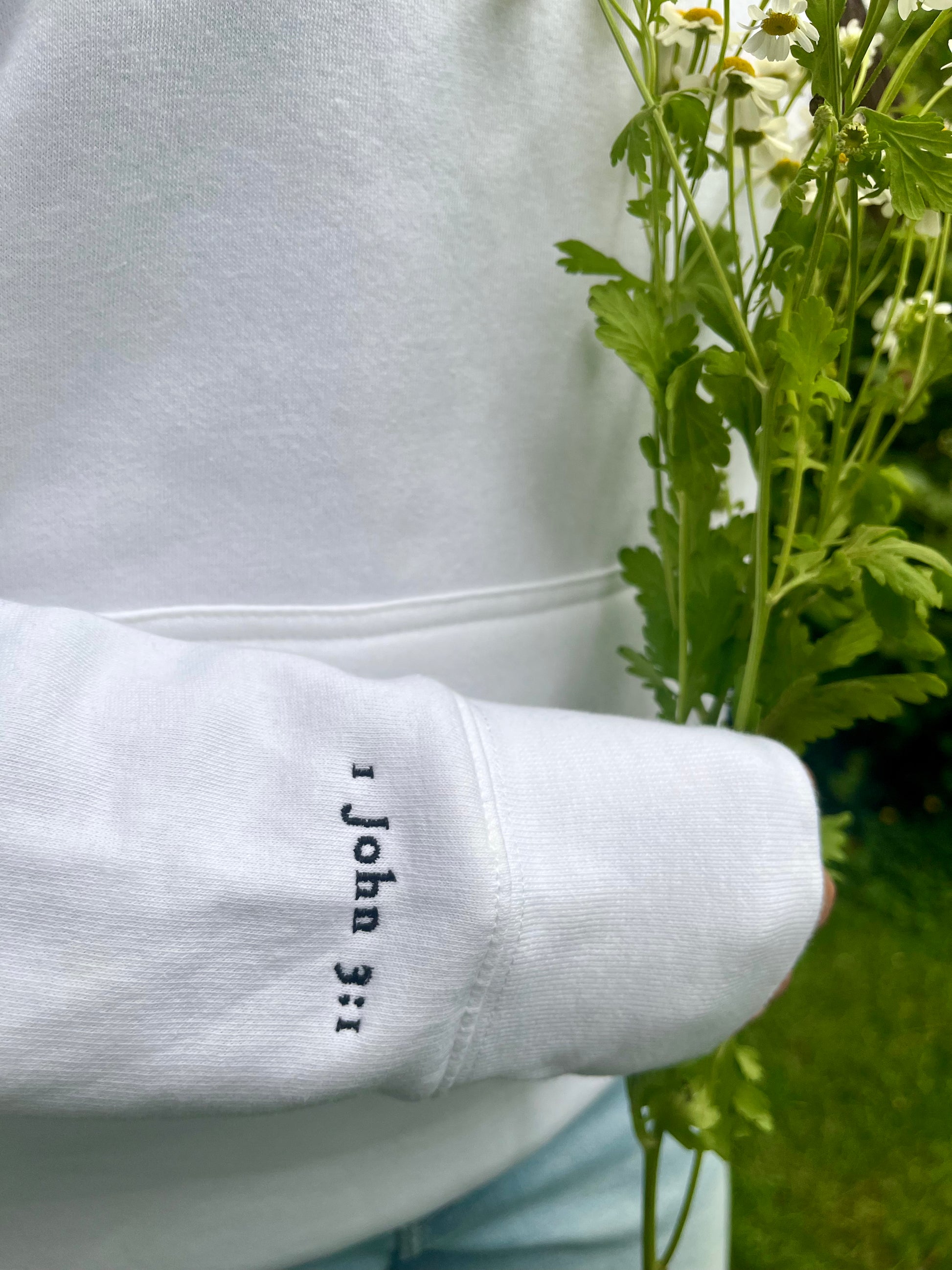 Detail of embroidered Bible verse “1 John 3:1” on white hoodie sleeve, held next to fresh flowers. Christian clothing with a purpose.