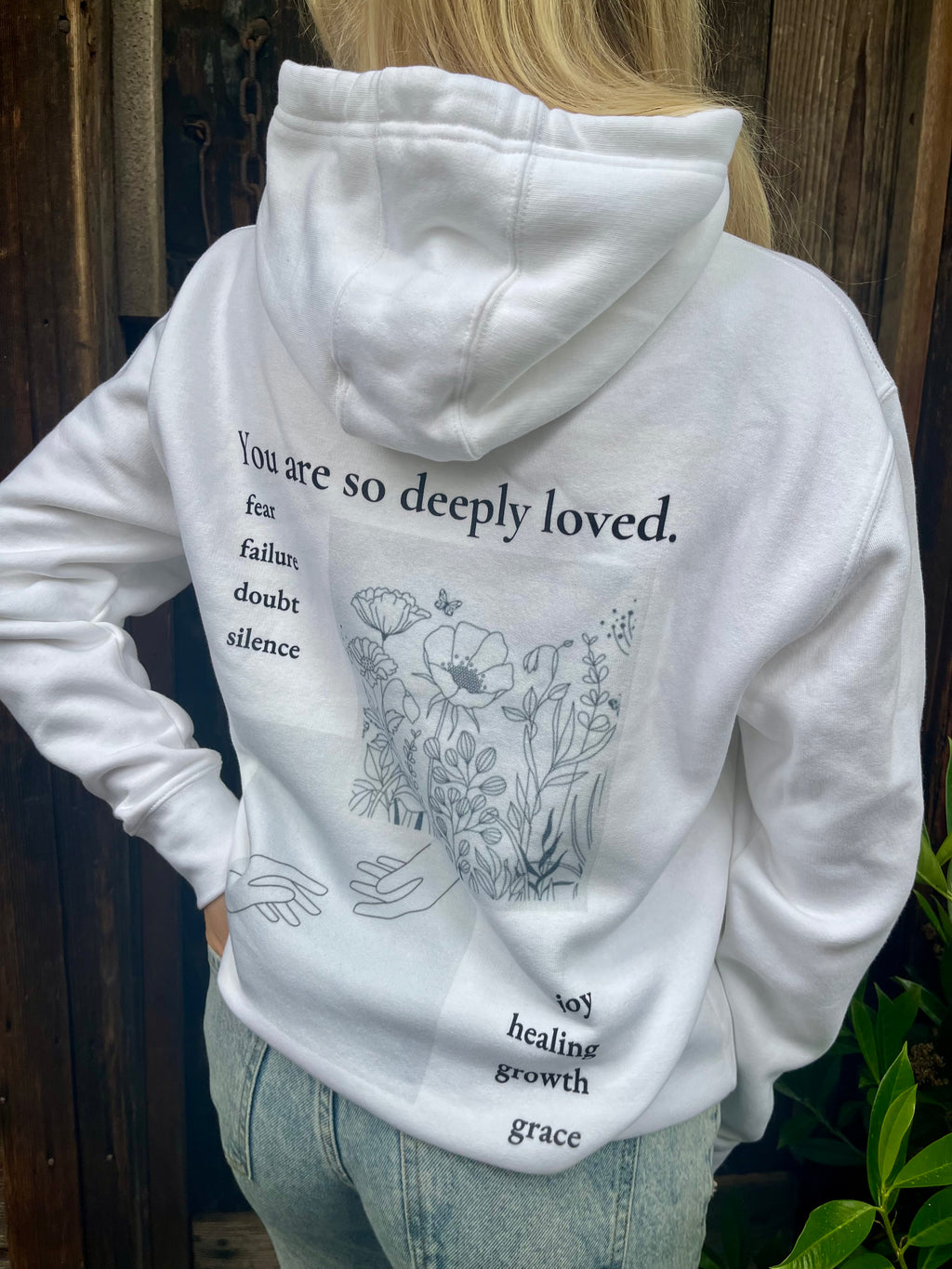 Back of white hoodie featuring hand-drawn floral artwork and the message “you are so deeply loved.” Encouraging words like fear, failure, and silence fade into healing, growth, and grace.