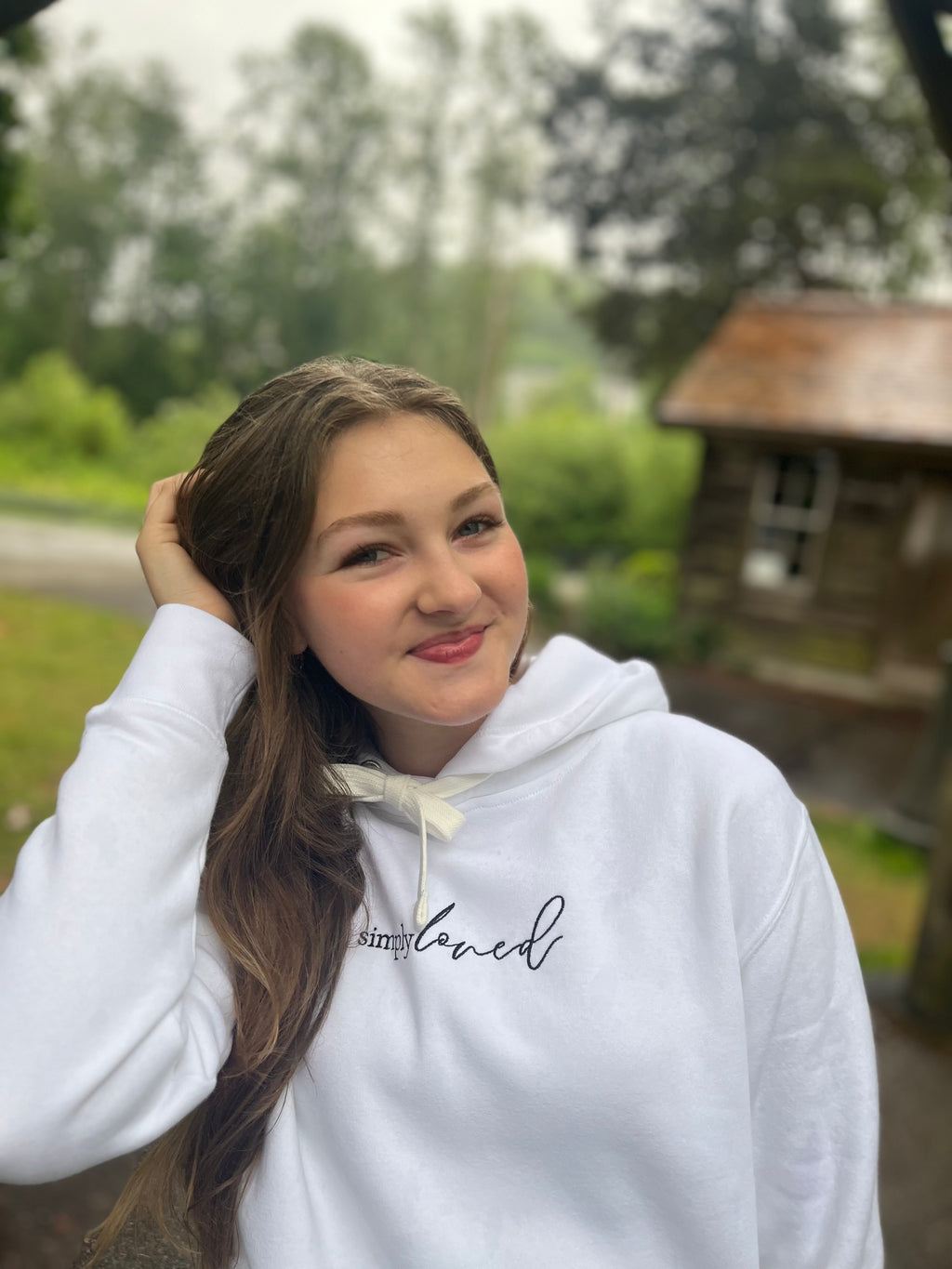 Girl in a “simply loved” hoodie smiling softly near a wooden cabin. Minimalist, scripture-rooted hoodie from Simply Sofia Joy.