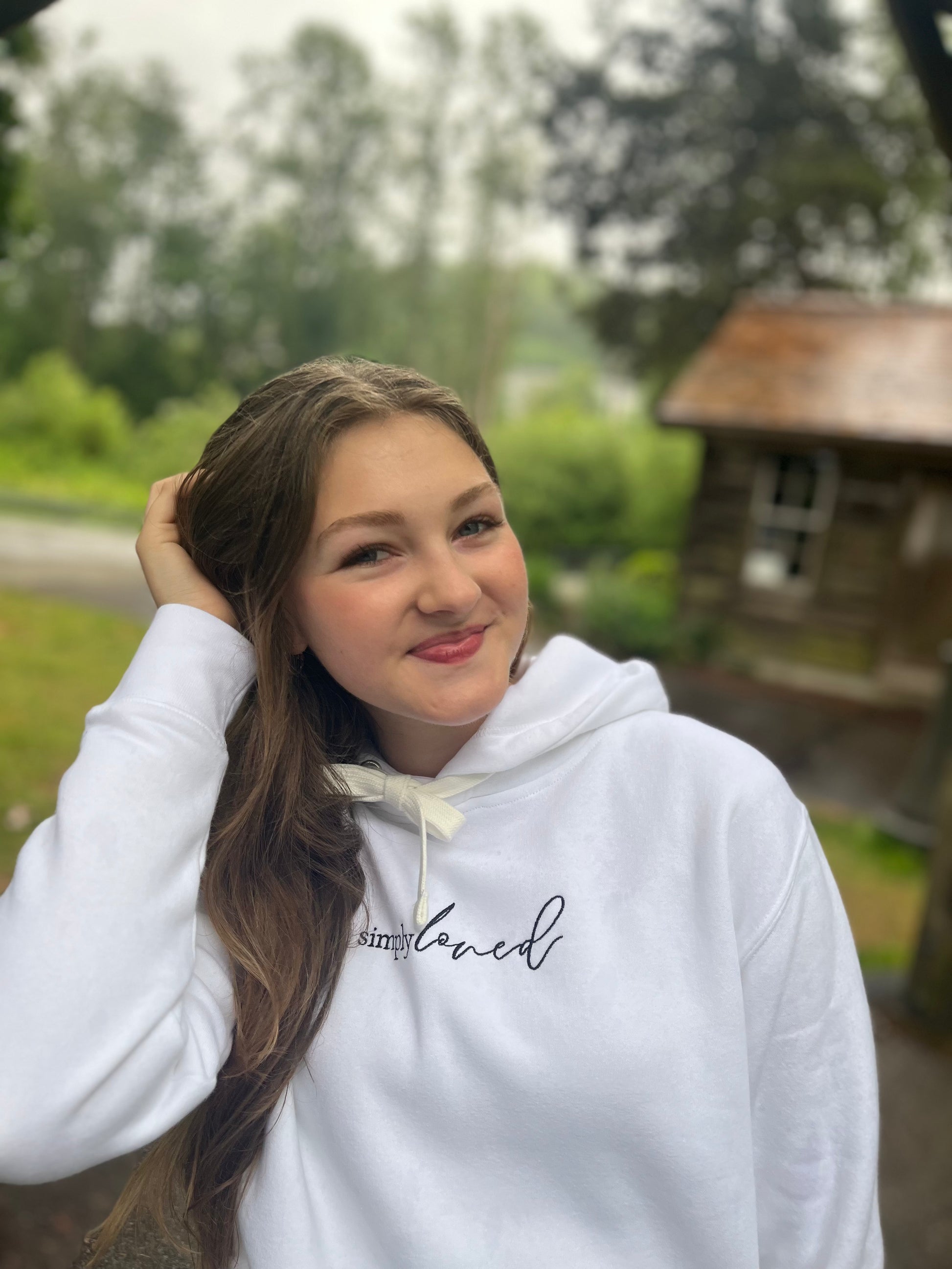 Girl in a “simply loved” hoodie smiling softly near a wooden cabin. Minimalist, scripture-rooted hoodie from Simply Sofia Joy.